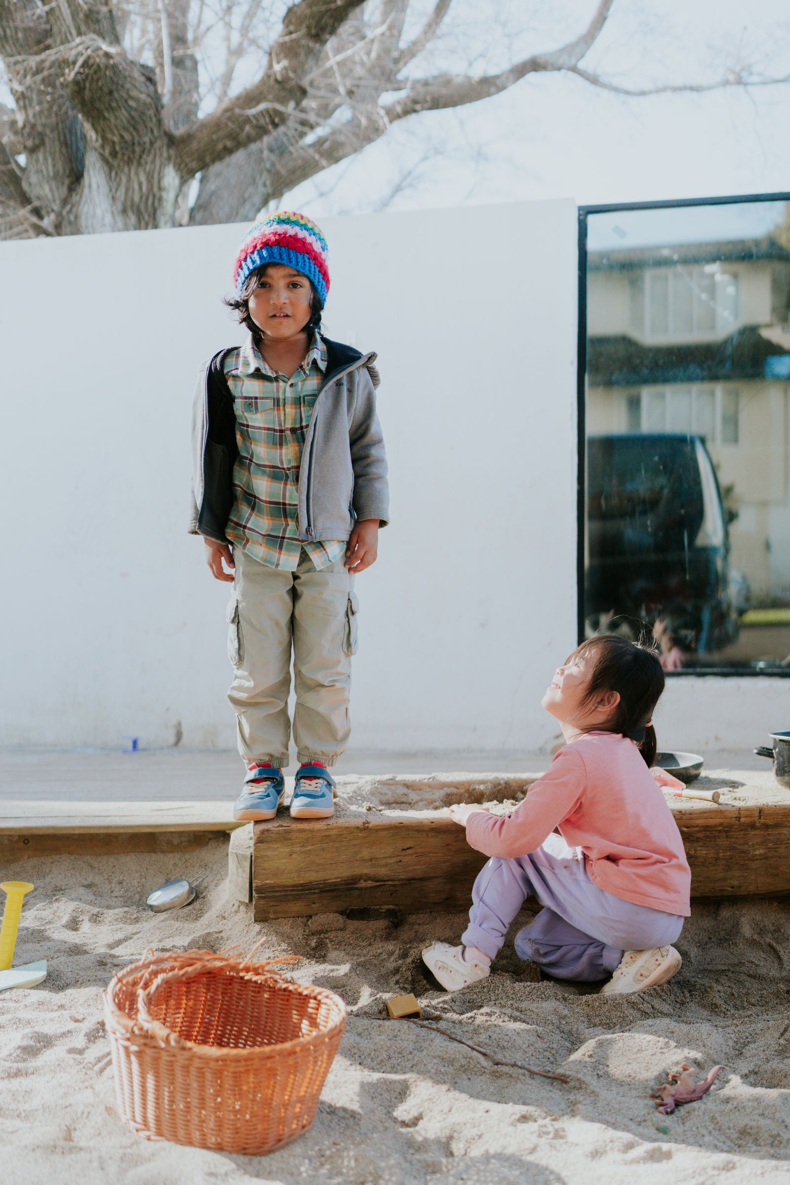A boy in a rainbow beanie stands at the edge of the sandpit