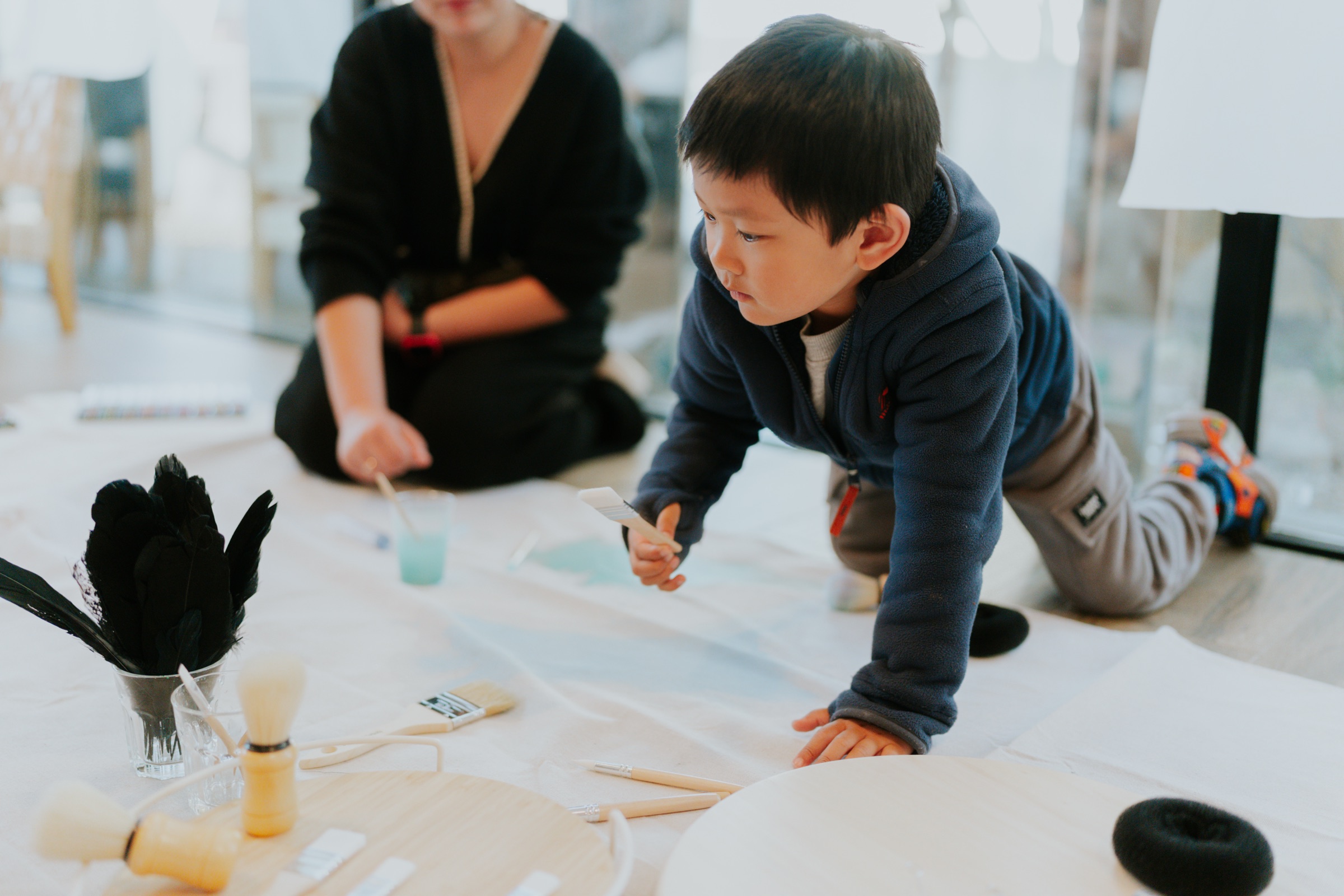 A young boy leans in to paint on paper laid across the floor
